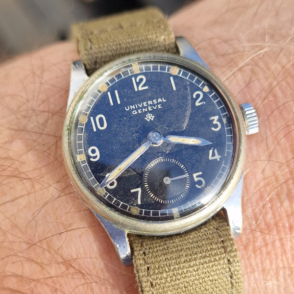 A close-up of a wrist wearing a vintage-style military watch with a blue dial, silver-tone case, and olive green canvas strap.
