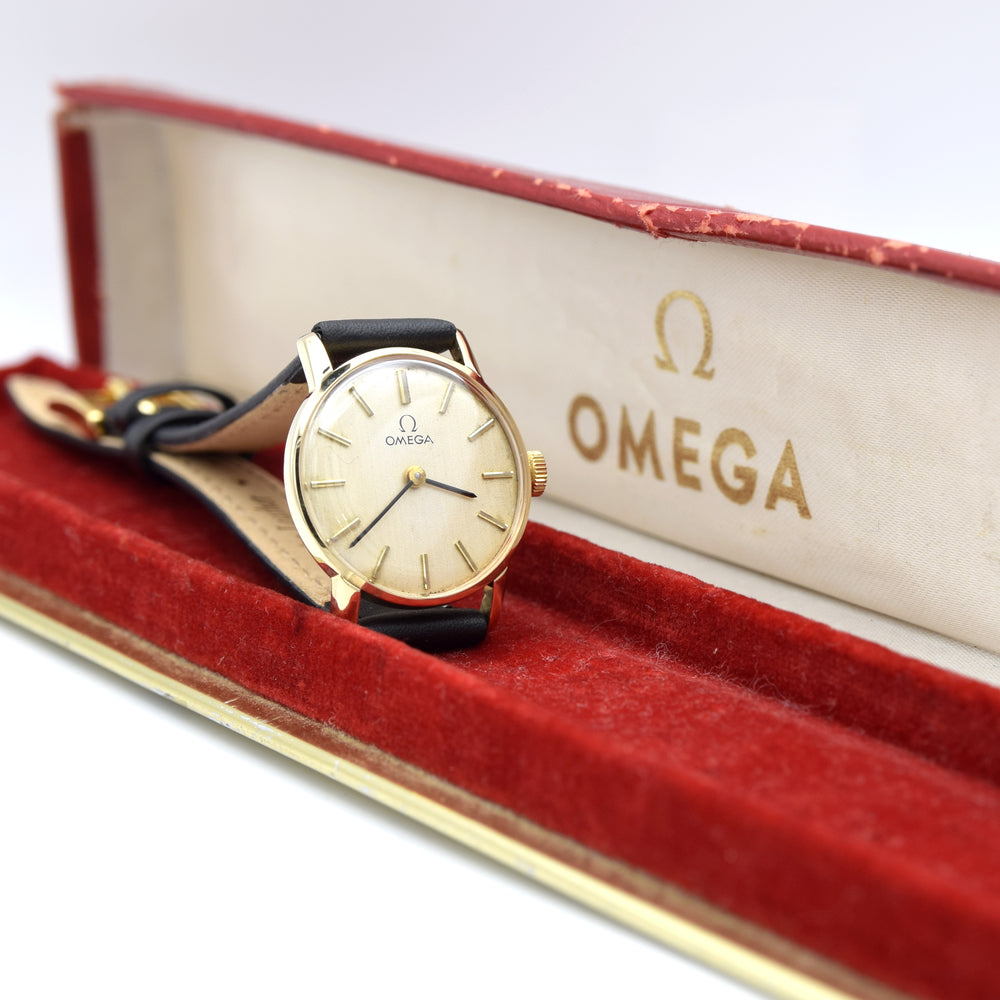 An Omega wristwatch with a gold case, cream dial, and black leather strap, displayed inside an open red velvet Omega presentation box.