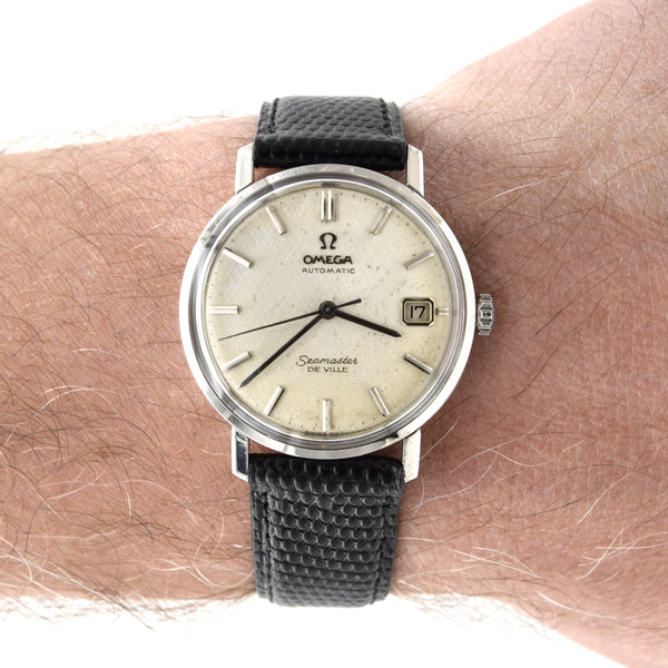 An Omega Seamaster De Ville wristwatch with a silver-toned case, cream-colored dial, and a black textured strap, shown worn on a person's wrist.