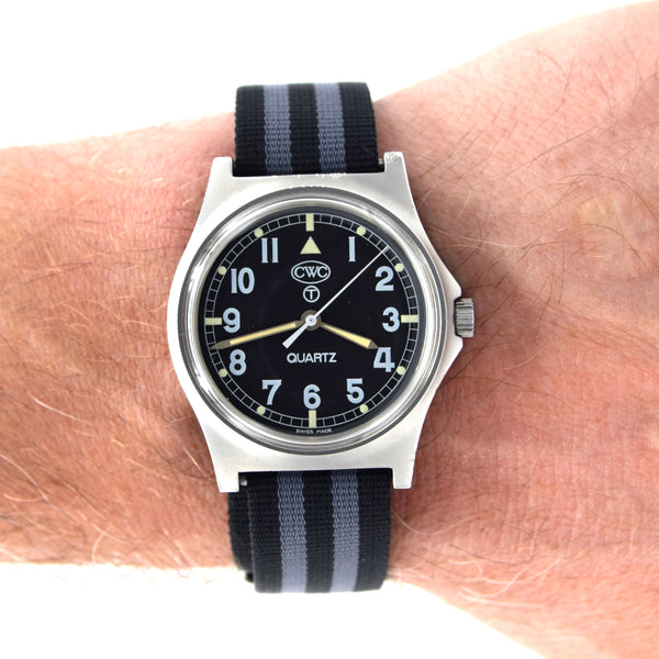 A wristwatch with a black dial, silver-toned case, and a black-and-gray striped fabric strap, shown worn on a person's wrist against a white background.