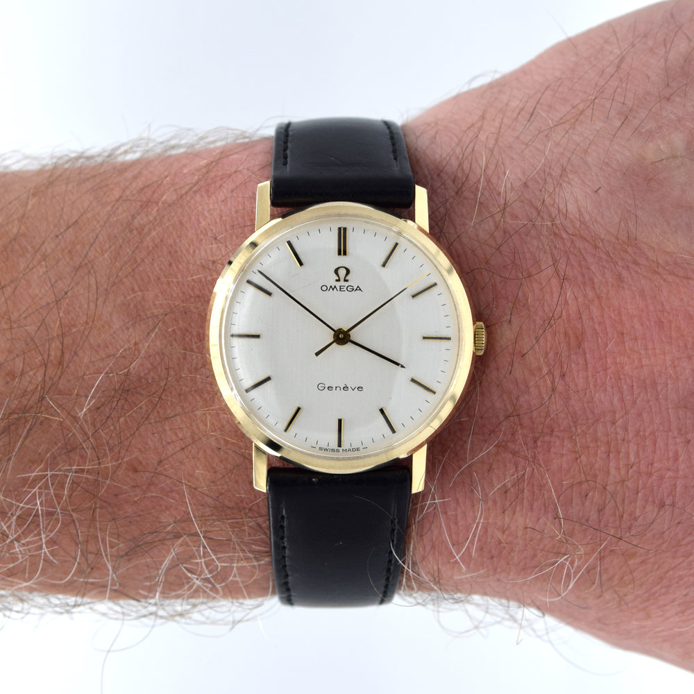 An Omega watch with a gold case, white dial, and black leather strap worn on a person's wrist against a white background.