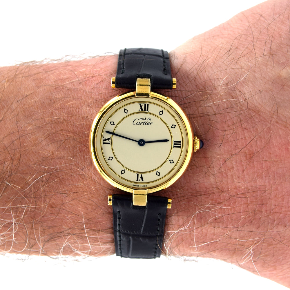 A Cartier watch with a gold case, cream dial, and black leather strap, shown worn on a person's wrist against a white background.