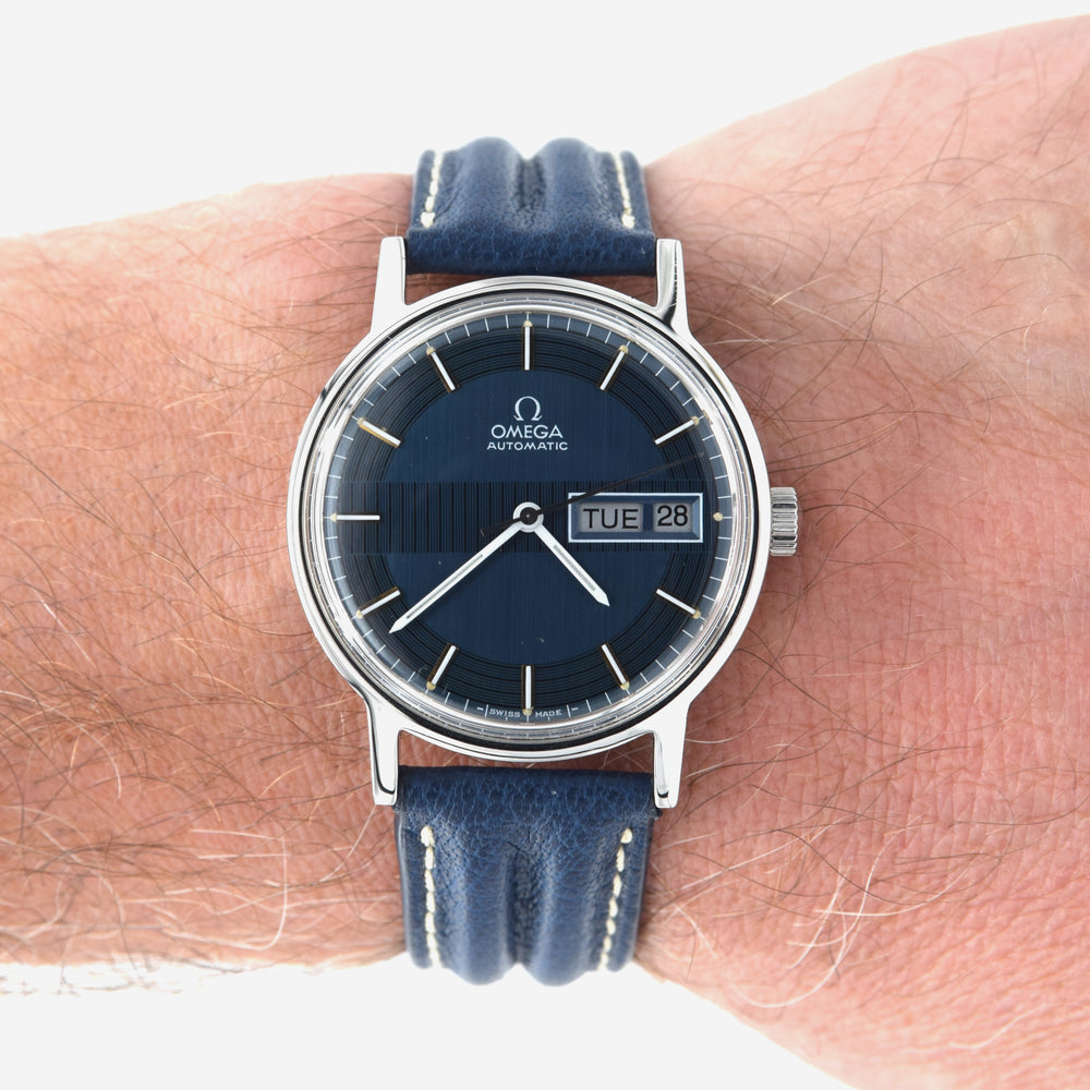 Omega Automatic wristwatch with a dark blue dial and blue leather strap, shown worn on a person's wrist against a white background.