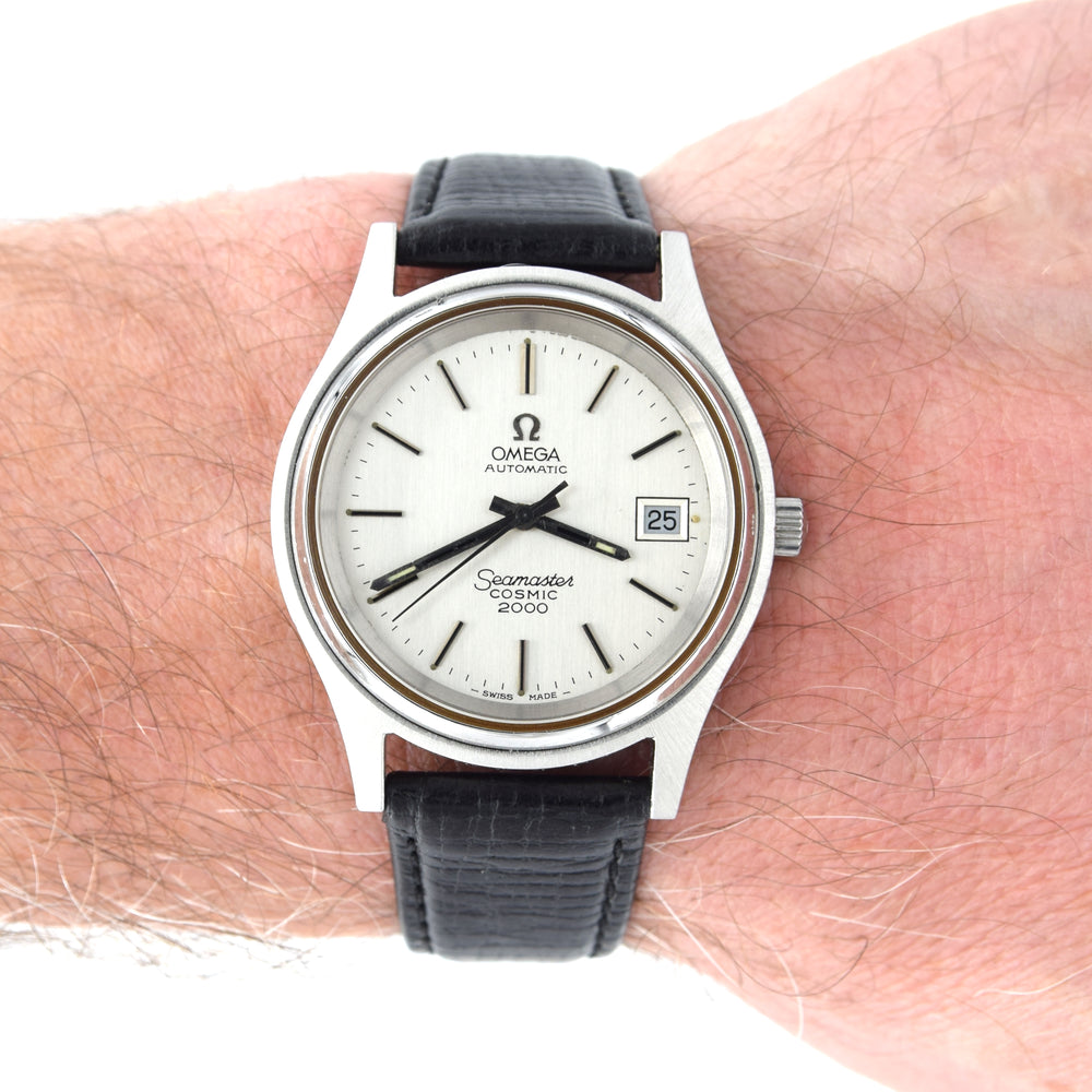 A vintage Omega Automatic cosmic 2000 watch with a silver-toned metal case, white dial, and black leather strap, shown worn on a person's wrist.