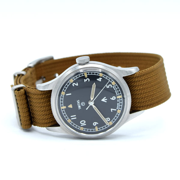 A vintage-style wristwatch with a brown canvas strap, a silver-toned metal case, and a dark dial featuring white Arabic numerals and a black tachymeter scale.