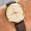 Wrist wearing a gold watch with a brown leather strap on a neutral background