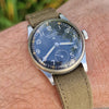 A wristwatch with a dark blue dial and olive green canvas strap, shown worn on a person's wrist outdoors with blurred green grass in the background.