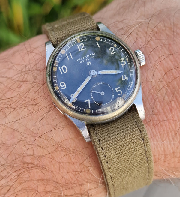 A wristwatch with a dark blue dial and olive green canvas strap, shown worn on a person's wrist outdoors with blurred green grass in the background.