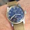 A close-up of a wrist wearing a vintage-style military watch with a blue dial, silver-tone case, and olive green canvas strap.