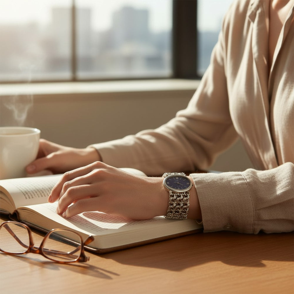 A close-up lifestyle shot of a person wearing a silver-toned metal link watch with a dark blue dial, sitting at a wooden desk with an open book, a steaming mug, and eyeglasses in a sunlit room.