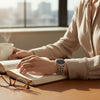 A close-up lifestyle shot of a person wearing a silver-toned metal link watch with a dark blue dial, sitting at a wooden desk with an open book, a steaming mug, and eyeglasses in a sunlit room.