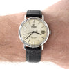 An Omega Seamaster De Ville wristwatch with a silver-toned case, cream-colored dial, and a black textured strap, shown worn on a person's wrist.