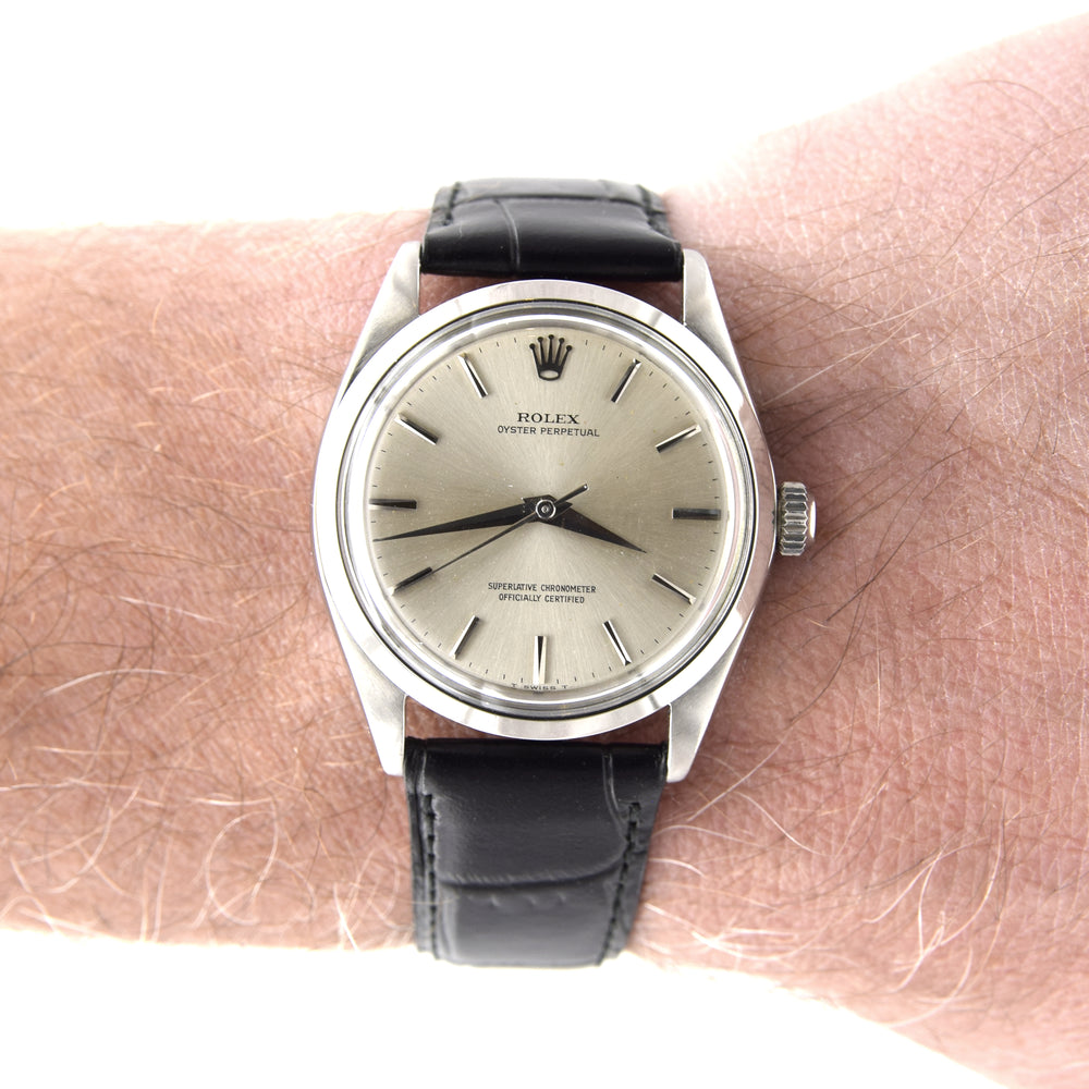 A vintage Rolex Oyster Perpetual watch with a silver-toned metal case, beige dial, and black leather strap, shown worn on a person's wrist against a white background.