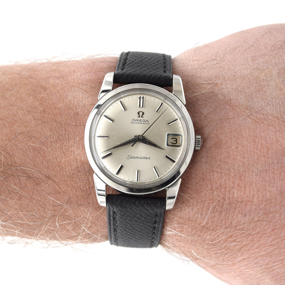 A vintage Omega Seamaster wristwatch with a silver-toned stainless steel case, a cream-colored dial, and a black textured leather strap, shown worn on a person's wrist.
