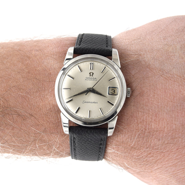 A vintage Omega Seamaster wristwatch with a silver-toned stainless steel case, a cream-colored dial, and a black textured leather strap, shown worn on a person's wrist.