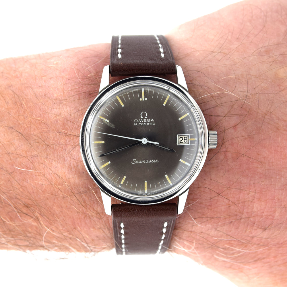 A wristwatch with a dark brown leather strap and a dark brown dial, worn on a person's wrist. The watch features a silver-tone stainless steel case and yellow-gold indices, with the Omega Seamaster logo visible on the dial.