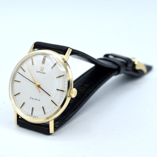 Omega Geneva watch with a gold-tone case, white dial, and black leather strap, photographed on a light blue-white background.