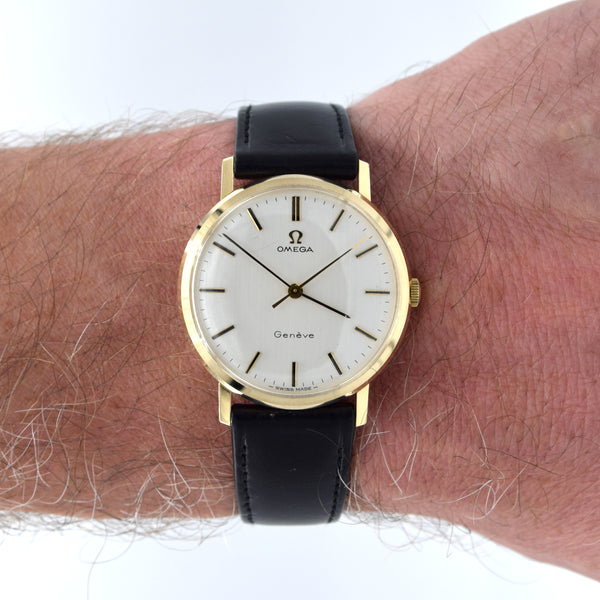 An Omega watch with a gold case, white dial, and black leather strap worn on a person's wrist against a white background.