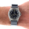 A wristwatch with a black dial, silver-toned metal case, and a dark gray fabric strap, shown worn on a person's wrist against a white background.
