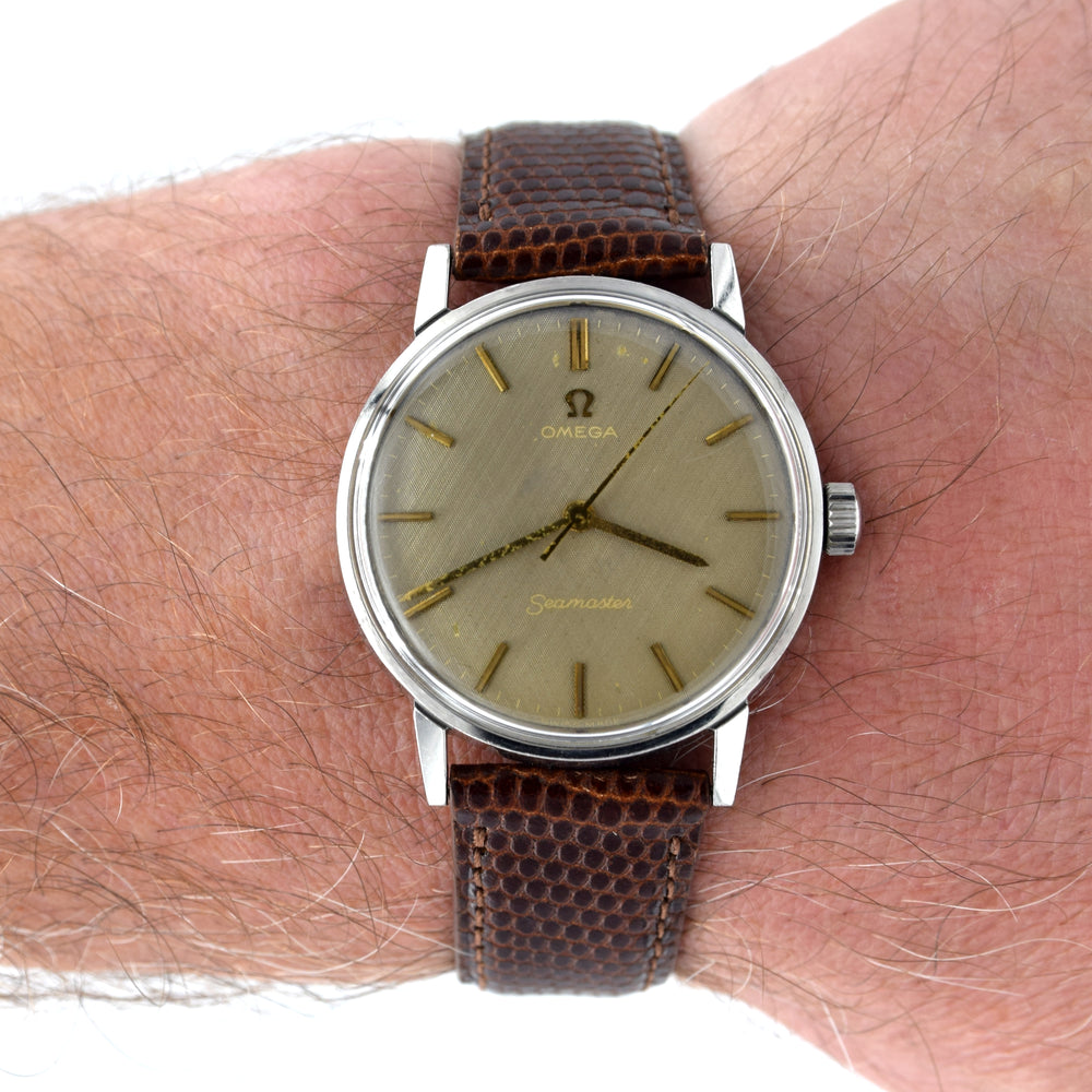 A vintage Omega Seamaster wristwatch with a brown textured leather strap and a champagne-gold dial, shown worn on a person's wrist against a white background.