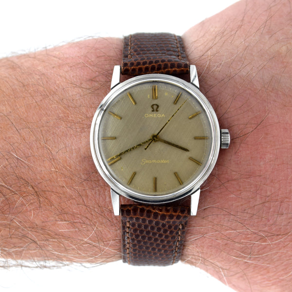 A vintage Omega Seamaster wristwatch with a brown textured leather strap and a champagne-gold dial, shown worn on a person's wrist against a white background.