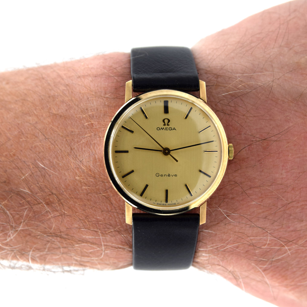 A vintage Omega Geneva watch with a gold dial and gold case, worn on a person's wrist. The watch features a black leather strap and black stick indices.