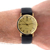 A vintage Omega Geneva watch with a gold dial and gold case, worn on a person's wrist. The watch features a black leather strap and black stick indices.