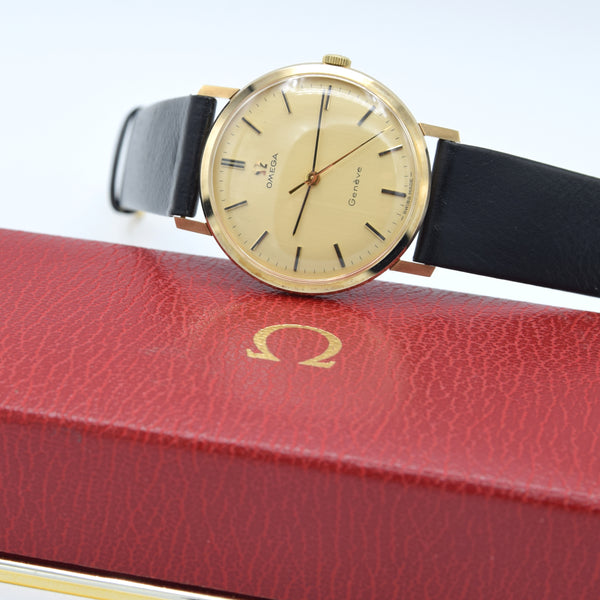 A vintage Omega  wristwatch with a gold-toned case, beige dial, and black leather strap, resting on a red textured leather Omega presentation box.