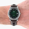 An Omega Automatic wristwatch with a dark green dial and a dark brown leather strap, shown worn on a person's wrist against a white background.
