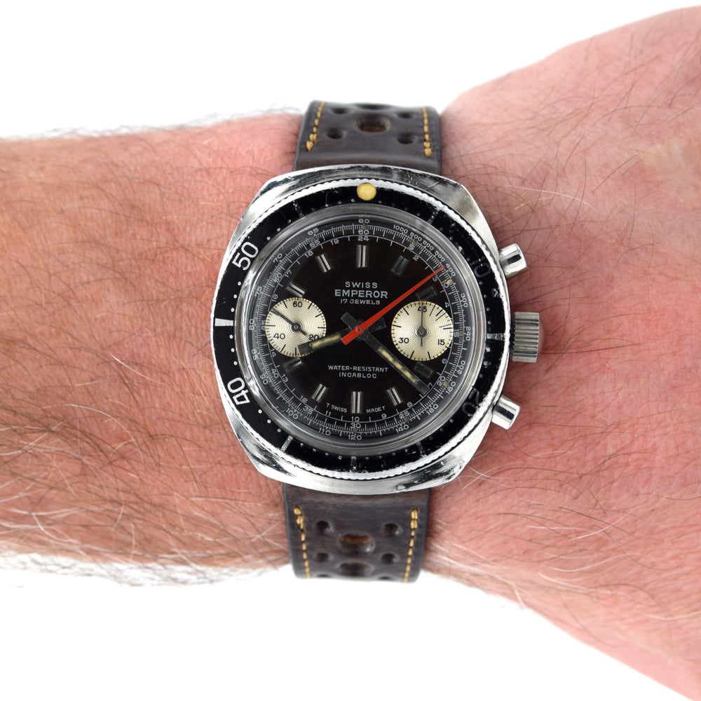 A vintage-style wristwatch with a silver-toned metal case, black dial, and perforated black strap with yellow stitching, shown worn on a person's wrist against a white background.