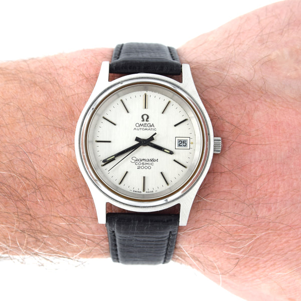 A vintage Omega Automatic cosmic 2000 watch with a silver-toned metal case, white dial, and black leather strap, shown worn on a person's wrist.