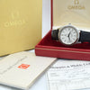 Omega wristwatch displayed in an open red presentation box, with the gold Omega box lid, booklet, and papers visible around it.