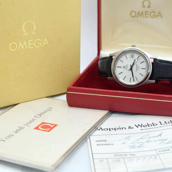 Omega wristwatch displayed in an open red presentation box, with the gold Omega box lid, booklet, and papers visible around it.
