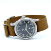 A vintage-style wristwatch with a brown canvas strap, a silver-toned metal case, and a dark dial featuring white Arabic numerals and a black tachymeter scale.
