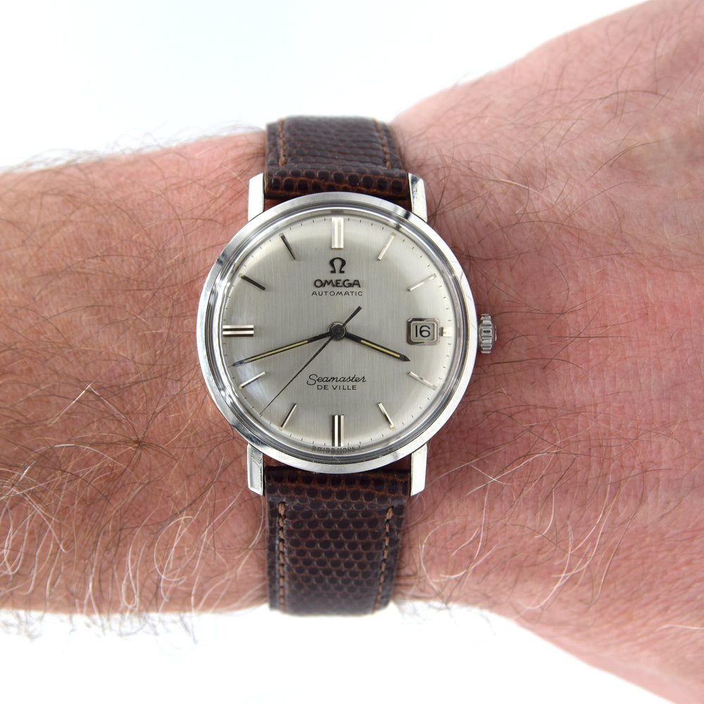 A vintage Omega De Ville watch with a silver dial and brown textured leather strap, shown worn on a person's wrist against a white background.