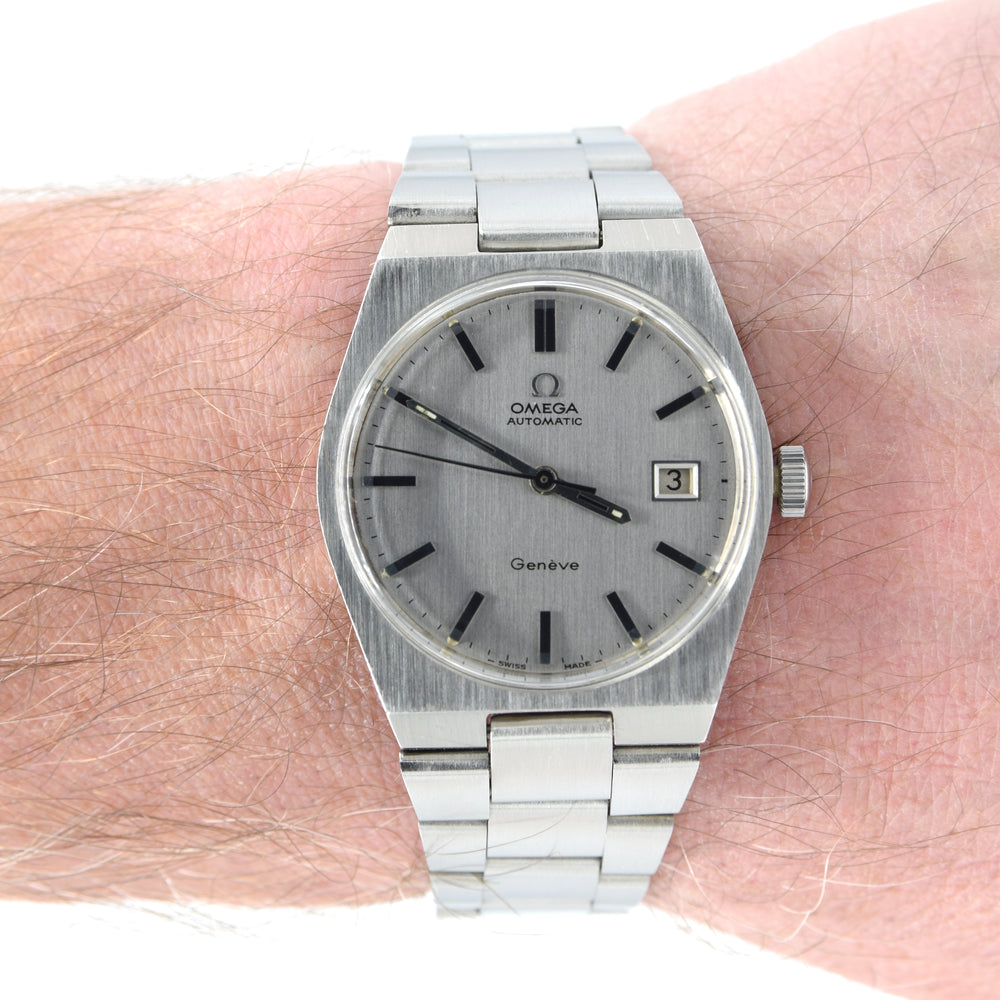 An Omega Automatic Geneve watch with a brushed silver stainless steel case and link bracelet, worn on a person's wrist. The watch features a silver dial with black stick indices and a date window at the 3 o'clock position.