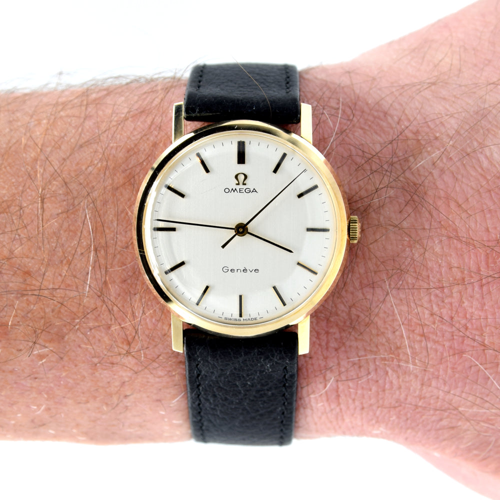 An Omega wristwatch with a gold-tone case, white dial, and black leather strap, shown worn on a person's wrist against a white background.