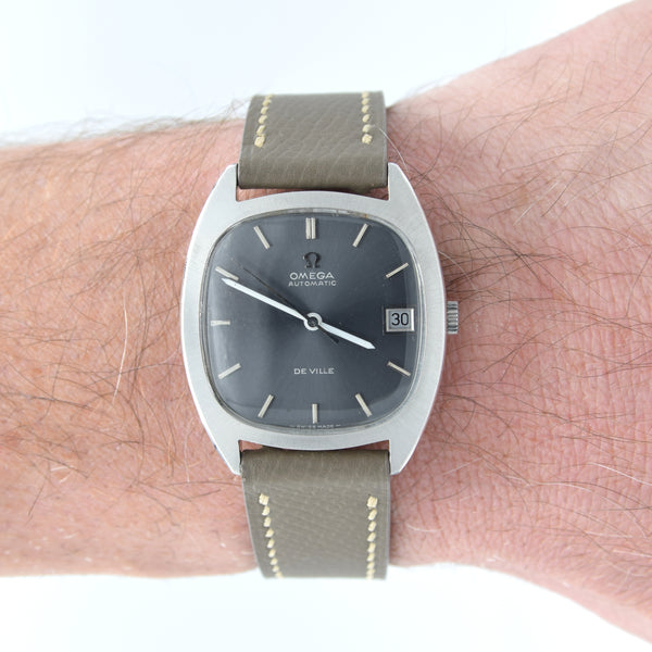 A vintage Omega De Ville wristwatch with a silver-toned case, dark gray dial, and olive green leather strap, shown worn on a person's wrist.